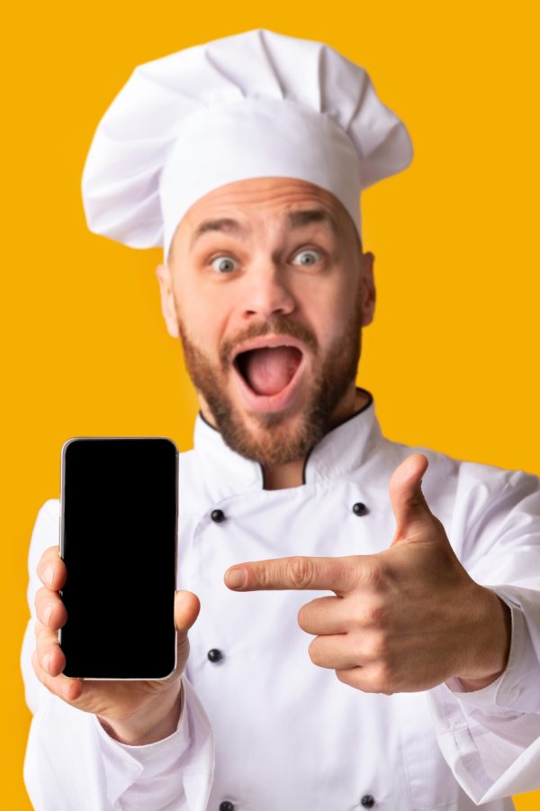 Food Delivery Application. Excited Chef Man Showing Smartphone Blank Screen Recommending Catering Service Posing Over Yellow Background. Studio Shot, Panorama